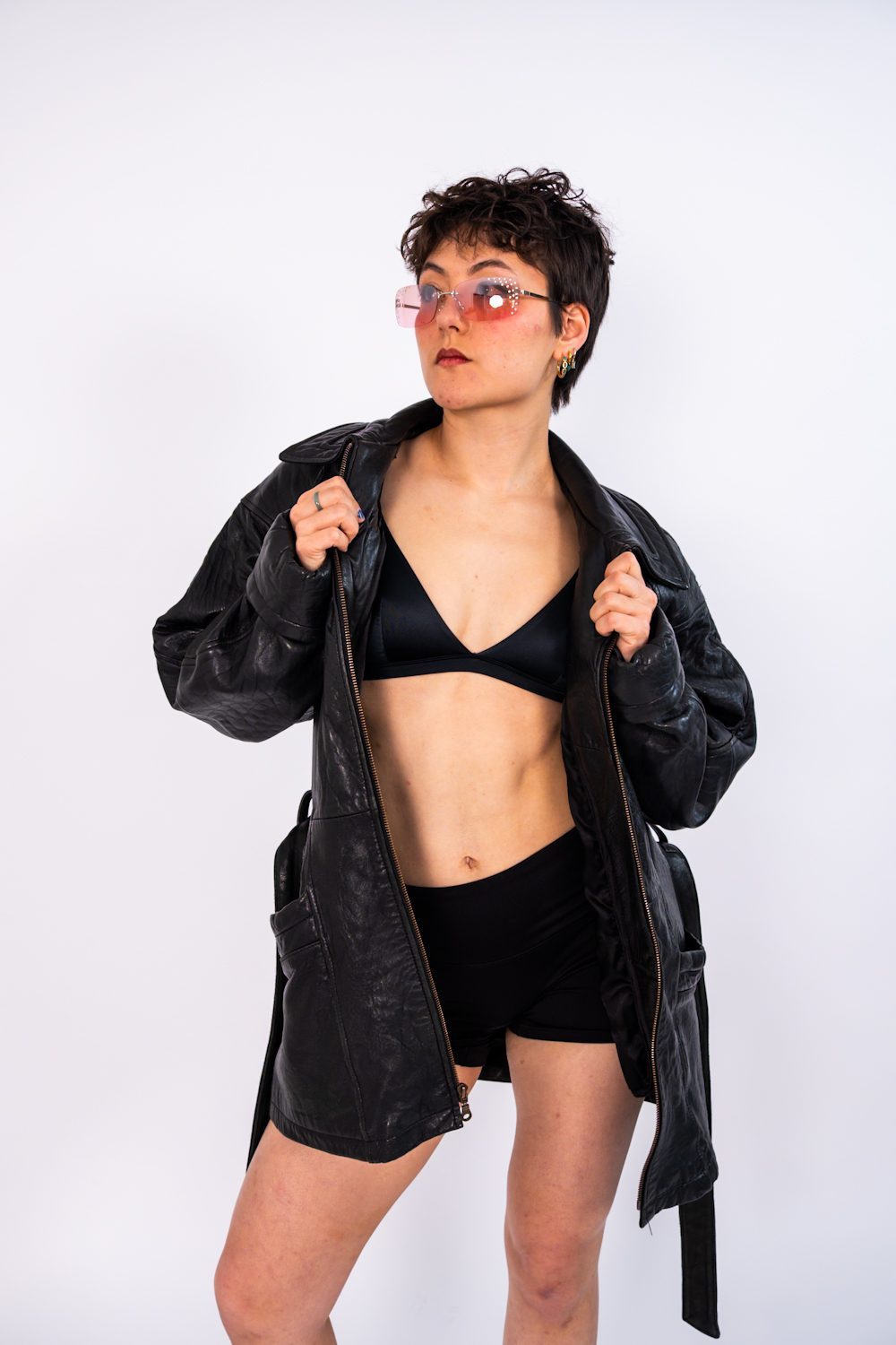 Full-length studio portrait on white, subject in an oversized leather jacket and pink tinted sunglasses.