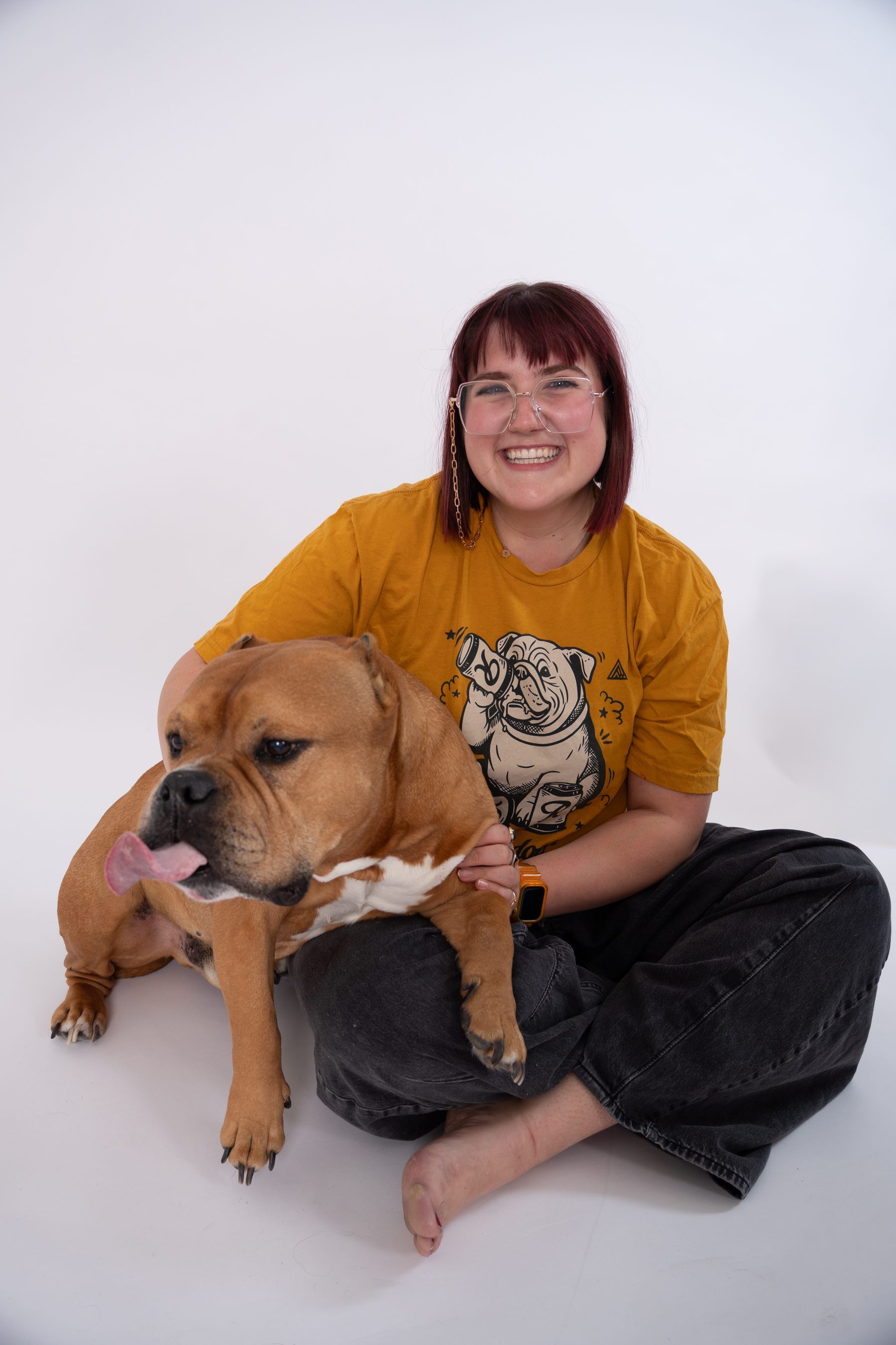 Studio portrait on white, subject sitting on the floor laughing with a bulldog in their lap.