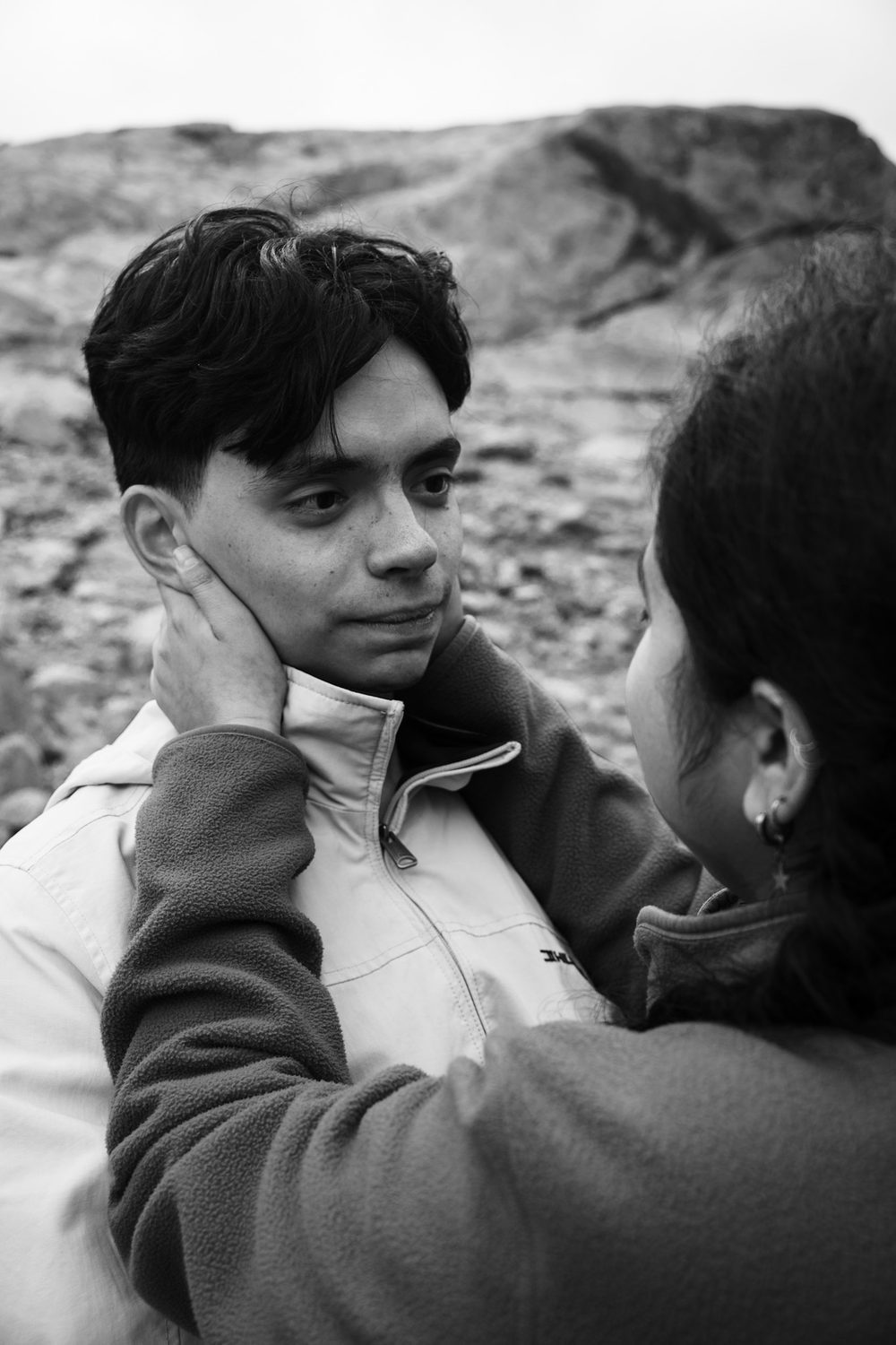 Black and white outdoor portrait, one person cupping the other's face, rocky hillside behind.