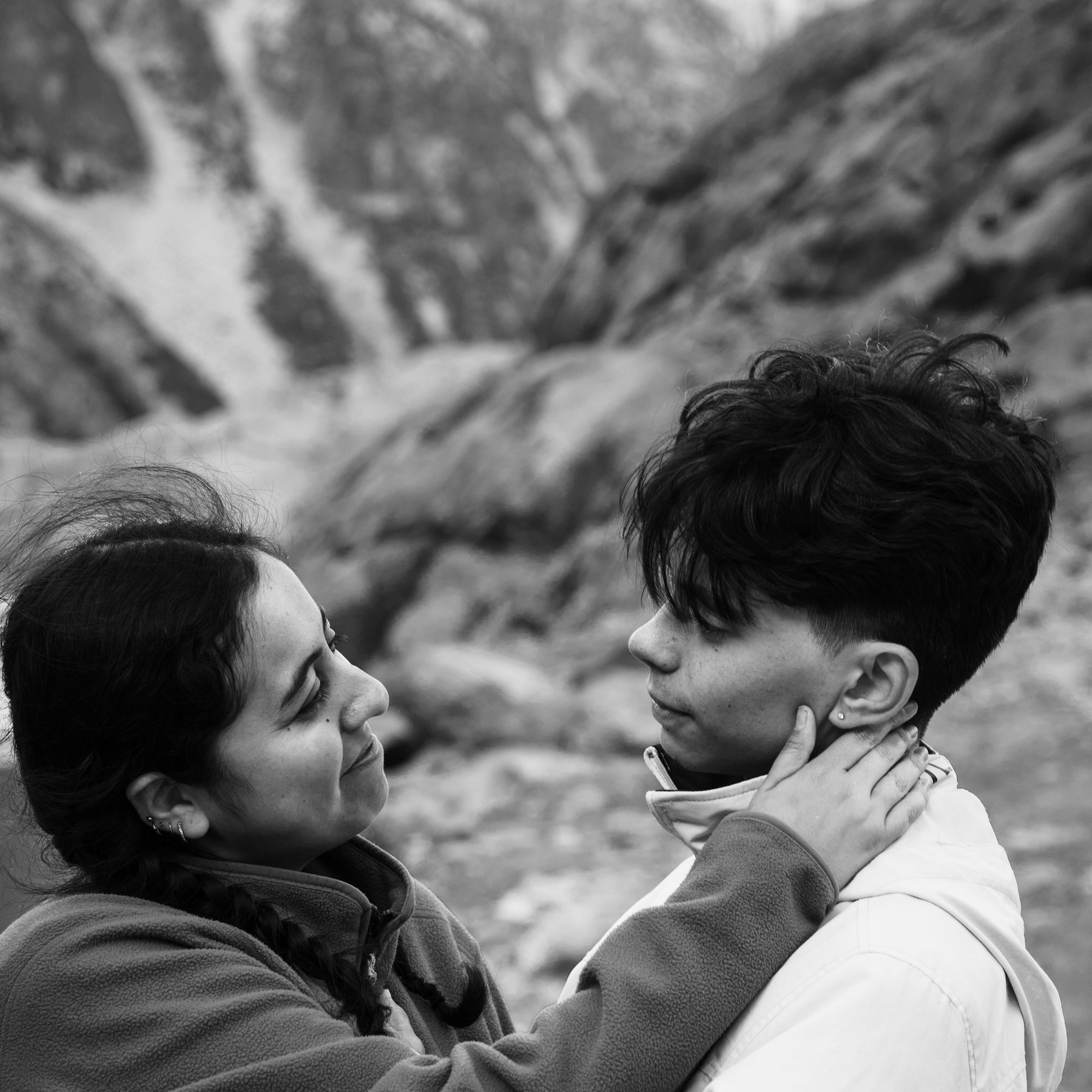 Black and white photograph of two people on a rocky shore, one holding the other's face.