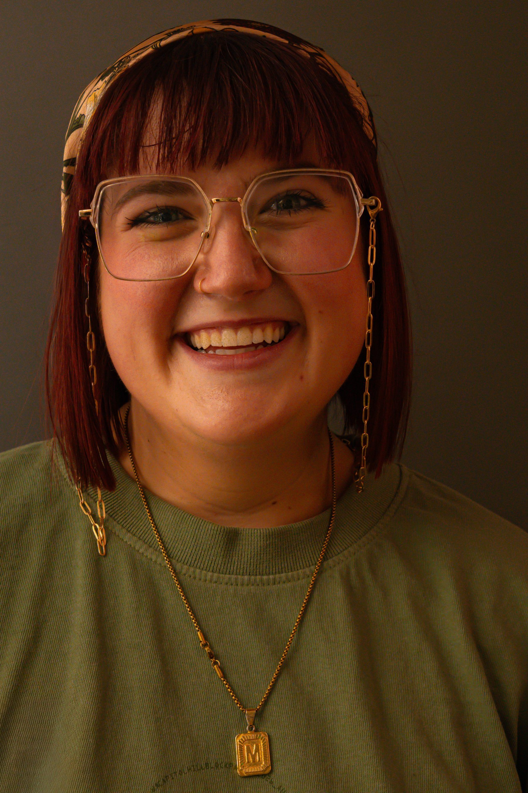 Studio portrait on an olive backdrop, subject with red hair and glasses laughing.
