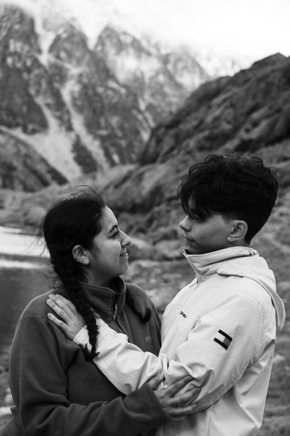 Black and white outdoor portrait of two people facing each other on a rocky shore, mountains behind.